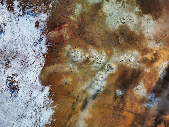 An aerial view of a geothermal landscape with steam vents and patches of white mineral deposits. The area is predominantly brown with various shades, interspersed with white and blue on one side, indicative of snow or frost. There are several circular formations suggesting volcanic activity.