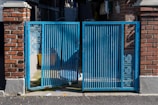 A blue metal gate with intricate geometric patterns stands between two brick pillars. The gate is partially open, revealing a shadowed area with a yellow container and other miscellaneous items. Sunlight casts shadows on the ground, highlighting the texture of the asphalt.