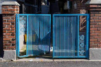 A blue metal gate with intricate geometric patterns stands between two brick pillars. The gate is partially open, revealing a shadowed area with a yellow container and other miscellaneous items. Sunlight casts shadows on the ground, highlighting the texture of the asphalt.