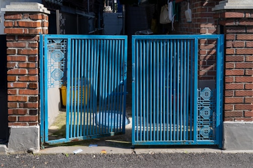 A blue metal gate with intricate geometric patterns stands between two brick pillars. The gate is partially open, revealing a shadowed area with a yellow container and other miscellaneous items. Sunlight casts shadows on the ground, highlighting the texture of the asphalt.