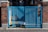A blue metal gate with intricate geometric patterns stands between two brick pillars. The gate is partially open, revealing a shadowed area with a yellow container and other miscellaneous items. Sunlight casts shadows on the ground, highlighting the texture of the asphalt.