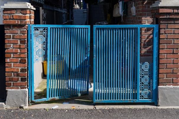 A blue metal gate with intricate geometric patterns stands between two brick pillars. The gate is partially open, revealing a shadowed area with a yellow container and other miscellaneous items. Sunlight casts shadows on the ground, highlighting the texture of the asphalt.