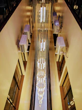 A vertical view down an indoor space with a tall, cascading chandelier made of lights and hanging cables. Air conditioning units are installed on the walls, and the light from the chandelier creates a warm glow against the beige walls.