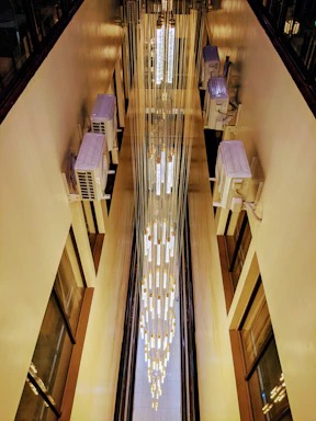 A vertical view down an indoor space with a tall, cascading chandelier made of lights and hanging cables. Air conditioning units are installed on the walls, and the light from the chandelier creates a warm glow against the beige walls.