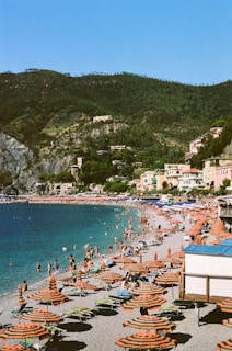 A bustling beach scene with numerous people enjoying the sun and water. The beach is lined with rows of orange and green striped umbrellas and matching sun loungers. Several people are swimming, walking, or lounging on the beach. The backdrop features green hills and a quaint town with colorful buildings, creating a picturesque coastal setting.