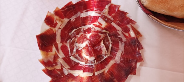 A plate artfully arranged with thin slices of cured ham in a circular pattern, showcasing a mixture of red and white hues. Next to the plate, there is a wicker basket with a piece of crusty bread.