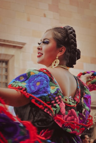 A person is wearing traditional colorful clothing featuring intricate floral embroidery and elaborate jewelry, with their hair styled in a braided updo. The background consists of a textured stone wall.