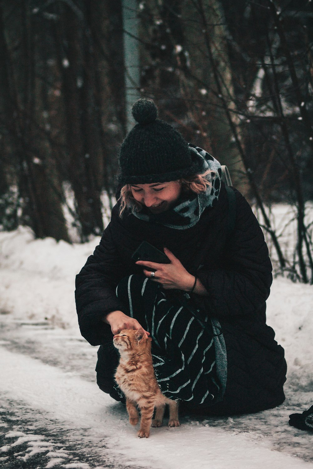a woman kneeling down next to a cat in the snow