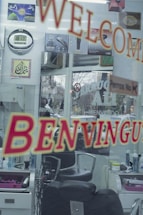 A hair salon interior seen through a glass door with welcoming signs in multiple languages. The reflection includes barber chairs, hair products, a wall clock, and decorative signage. Various posters and products are displayed on the walls.