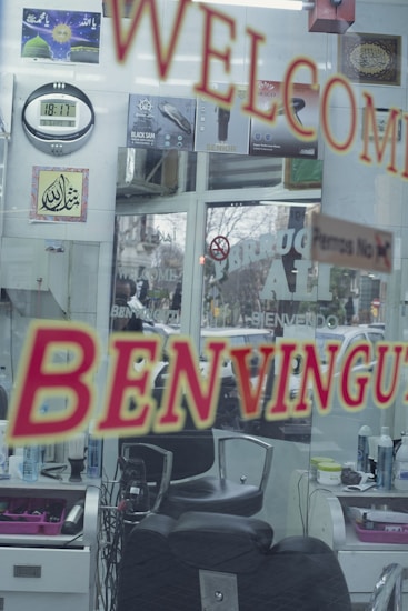 A hair salon interior seen through a glass door with welcoming signs in multiple languages. The reflection includes barber chairs, hair products, a wall clock, and decorative signage. Various posters and products are displayed on the walls.