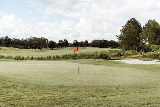 A serene golf course with an expansive green, featuring a centrally placed flagstick with a red flag. Surrounding the green are neatly trimmed grass areas, a sand bunker to the right, and a backdrop of lush green trees under a partly cloudy sky.