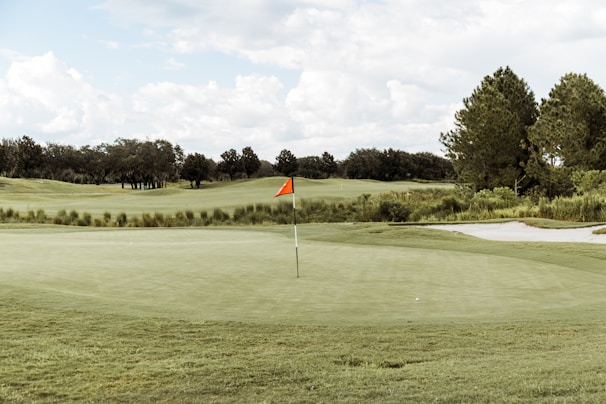 A serene golf course with an expansive green, featuring a centrally placed flagstick with a red flag. Surrounding the green are neatly trimmed grass areas, a sand bunker to the right, and a backdrop of lush green trees under a partly cloudy sky.