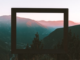 A mountain landscape is viewed through a large rectangular frame, with the setting or rising sun casting a warm glow on the peaks. Pine trees are visible in the foreground, and the scene conveys a sense of serene natural beauty.