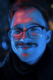 A person with glasses and a mustache, highlighted by a mix of blue and red lighting. The lighting creates dramatic shadows and gives an artistic, expressive feel. The person appears to be wearing a dark scarf or coat.