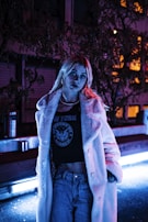 A person stands in an urban nighttime setting, illuminated by neon lights. The individual wears a long, white faux fur coat, a shirt with New York and an emblem printed on it, and jeans. The lighting casts a moody, purple and blue glow, highlighting the urban environment around them.