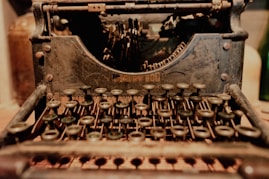 A close-up view of an antique, rusty typewriter with numerous keys visible in the foreground. The metallic body appears aged, with signs of wear and patina, and intricate mechanical parts are visible at the back. The lighting is warm, giving a nostalgic feel to the scene.