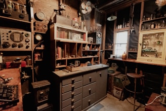 A cluttered workshop space features vintage electronics, books, and various tools. Shelves are filled with old equipment and bottles. A central cabinet with drawers is visible underneath more shelves containing old books and artifacts. The atmosphere is dimly lit with a vintage aesthetic.