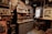A cluttered workshop space features vintage electronics, books, and various tools. Shelves are filled with old equipment and bottles. A central cabinet with drawers is visible underneath more shelves containing old books and artifacts. The atmosphere is dimly lit with a vintage aesthetic.
