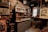 A cluttered workshop space features vintage electronics, books, and various tools. Shelves are filled with old equipment and bottles. A central cabinet with drawers is visible underneath more shelves containing old books and artifacts. The atmosphere is dimly lit with a vintage aesthetic.