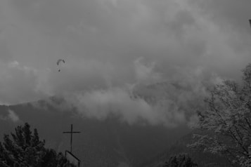 A grayscale image featuring a mountainous landscape shrouded in thick clouds. In the foreground, branches of trees are visible, some covered with snow. To the left, a cross stands atop a structure. A paraglider is visible in the sky, adding a sense of adventure to the scene.