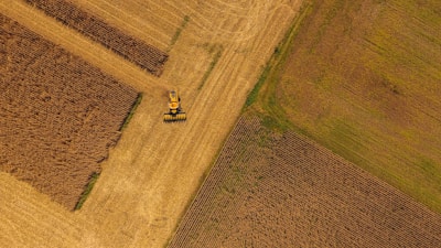 Aerial view of a vast agricultural landscape with neatly divided fields of various crops. A lone yellow tractor is working on one of the fields, creating a pattern in the earth. The fields are in shades of brown and yellow, indicating the harvesting season.