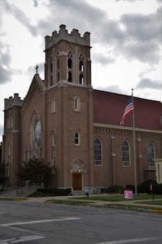 A large brick church building with a tall bell tower featuring arched windows and intricate architectural details. An American flag is raised on a pole in front of the church, adjacent to a small sign on the lawn. The sky is overcast, providing a subdued lighting to the scene.