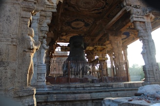 A stone structure featuring intricately carved pillars and statues surrounds a large, black stone sculpture enclosed by a metal fence. The ceiling displays ornate decorative patterns, and the area is in partial sunlight.