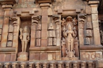 A detailed stone carving on an ancient structure features multiple figures, including a prominently displayed deity in the central niche with intricate details. The surrounding carvings include smaller figures and decorative elements, showcasing traditional artistic techniques.