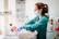 A healthcare professional wearing a mask and gloves is attending to a newborn in a hospital setting. The room contains medical equipment such as monitors and gauges. The newborn is lying in a bassinet, wrapped in a blanket.