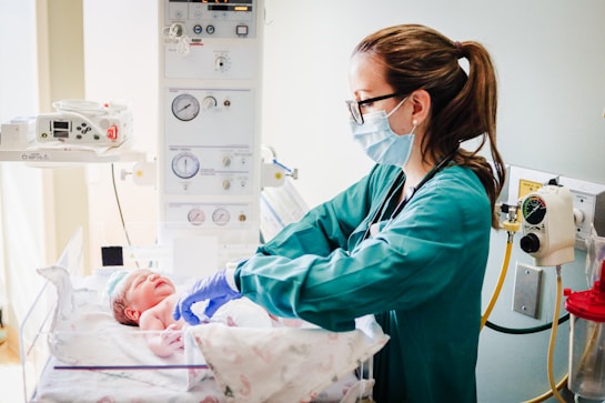A healthcare professional wearing a mask and gloves is attending to a newborn in a hospital setting. The room contains medical equipment such as monitors and gauges. The newborn is lying in a bassinet, wrapped in a blanket.
