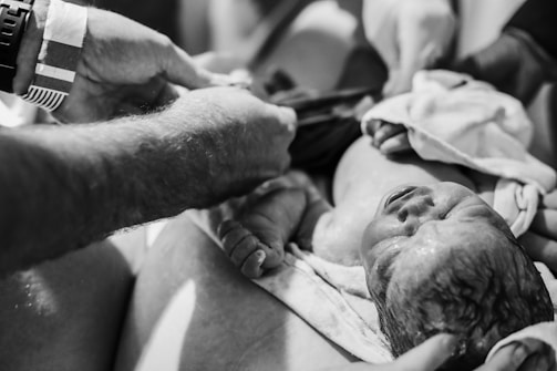 A black and white image of a newborn baby being held by several hands, possibly in a hospital or birthing setting. The baby is lying on a cloth, with one hand supporting its head. The image captures an intimate and raw moment shortly after birth.