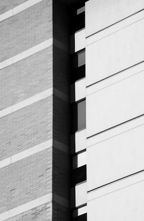 The image features a close-up view of a building's exterior, showcasing a combination of textured brick and smooth concrete surfaces. Vertical and horizontal lines create a geometric pattern, with windows intermittently breaking up the structure. The image is in black and white, emphasizing the contrast between light and shadow.