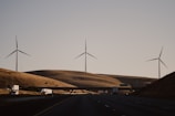 Wind turbines stand on rolling hills under a clear sky, with a highway and vehicles in the foreground. The scene exudes a sense of quiet efficiency and modern infrastructure amidst a natural landscape.