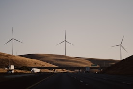 Wind turbines stand on rolling hills under a clear sky, with a highway and vehicles in the foreground. The scene exudes a sense of quiet efficiency and modern infrastructure amidst a natural landscape.