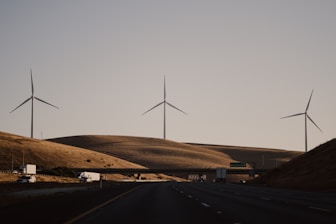 Wind turbines stand on rolling hills under a clear sky, with a highway and vehicles in the foreground. The scene exudes a sense of quiet efficiency and modern infrastructure amidst a natural landscape.