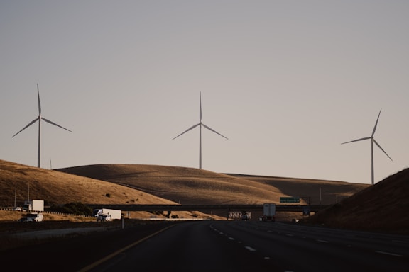 Wind turbines stand on rolling hills under a clear sky, with a highway and vehicles in the foreground. The scene exudes a sense of quiet efficiency and modern infrastructure amidst a natural landscape.