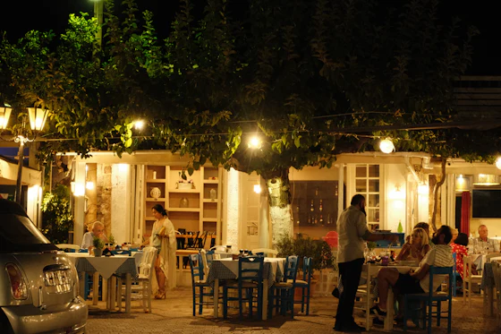 Blue chairs and laid tables under a tree strung with lit bulbs outside a taverna after dark, a waiter working between them.