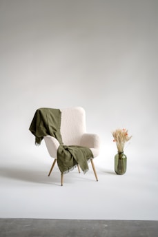 A mid-century modern armchair with a light fabric is draped with a green throw. Beside the chair, there is a green glass vase filled with dried flowers and grasses. The setting is minimalist with a plain background.