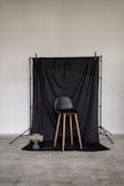 A minimalist setup featuring a black chair with wooden legs positioned in front of a black draped backdrop. The backdrop is supported by a stand, and a small transparent vase with white flowers is placed on the floor to the left of the chair. The surrounding environment includes a plain, light-colored wall and a concrete floor.