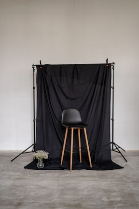 A minimalist setup featuring a black chair with wooden legs positioned in front of a black draped backdrop. The backdrop is supported by a stand, and a small transparent vase with white flowers is placed on the floor to the left of the chair. The surrounding environment includes a plain, light-colored wall and a concrete floor.