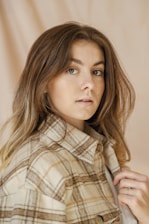 A person with long brown hair and neutral expression is wearing a plaid jacket. The background is a soft beige color that complements the overall earthy tones.