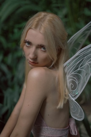 A person with long blonde hair is dressed as a fairy with translucent wings. Surrounding them are lush green plants, creating a mystical forest-like atmosphere.