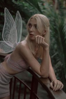 A person with blonde hair is leaning over a wooden railing in a lush, green environment. They are wearing a light-colored, strapless outfit and have transparent fairy wings. Their expression is contemplative, with a slightly open mouth and their hand resting near their chin.