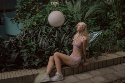 A woman with long blonde hair and fairy wings sits on the edge of a planter surrounded by lush green foliage. She wears a light pink two-piece outfit and white sneakers. The setting appears to be an indoor garden with tropical plants.