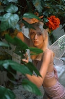 A person with long blonde hair surrounded by lush green leaves. They are wearing a sleeveless outfit in a soft pink color that sparkles. A red flower hangs nearby, and delicate fairy wings are visible on their back, adding a whimsical touch. The setting appears to be an outdoor garden or natural environment.