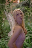 A person with long blond hair is seen amidst lush green foliage, wearing delicate fairy-like wings and a shimmery top, looking upward.