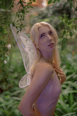A person with long blond hair is seen amidst lush green foliage, wearing delicate fairy-like wings and a shimmery top, looking upward.