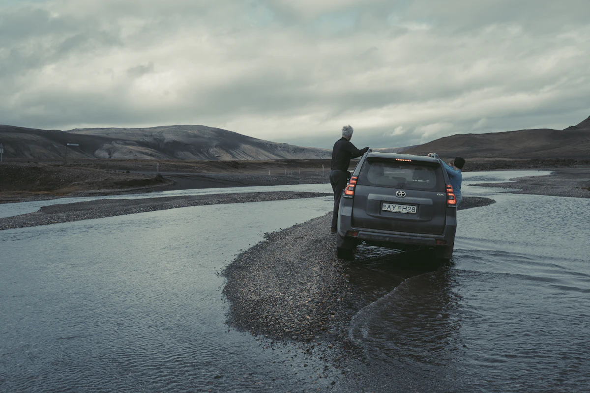 A four-wheel drive stopped in a wide, shallow river on a highland track, under gray cloud