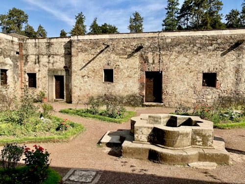 A stone courtyard featuring a weathered stone wall with wooden doors and small windows. The garden is lined with lush greenery and vibrant flowers. A centrally positioned, geometric stone structure adds an element of symmetry. Tall trees frame the scene under a clear blue sky.