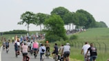 A large group of people is cycling together on a rural road, surrounded by green fields and trees. The cyclists are wearing casual clothing, and some are equipped with helmets. The road is slightly winding and appears to be part of a peaceful countryside setting.